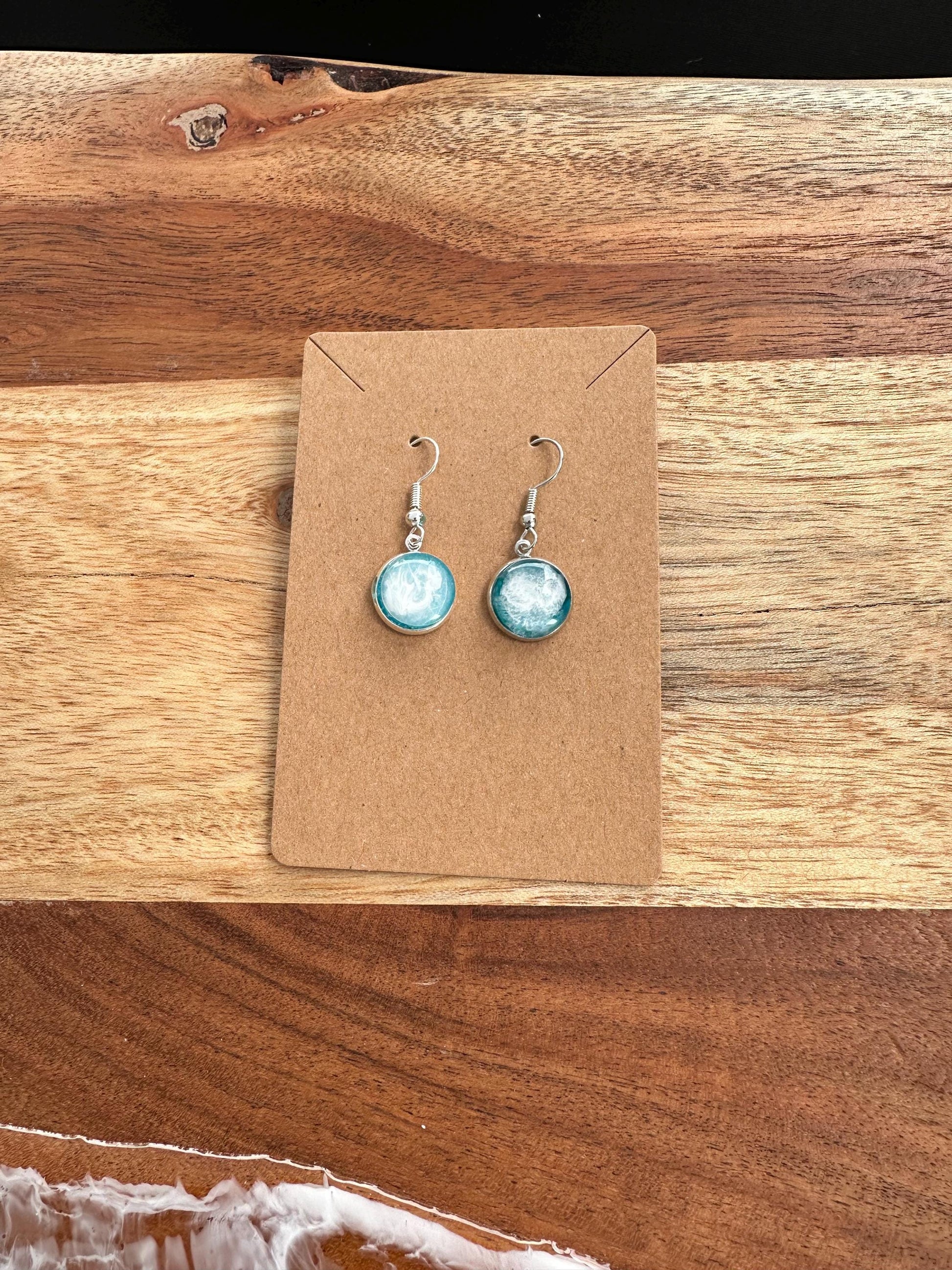 A pair of resin earrings featuring swirling shades of blue and green, resembling a stormy sea. The earrings are glossy, with silver hooks and a unique wave pattern in each piece. Set against a neutral background to highlight the ocean-inspired design