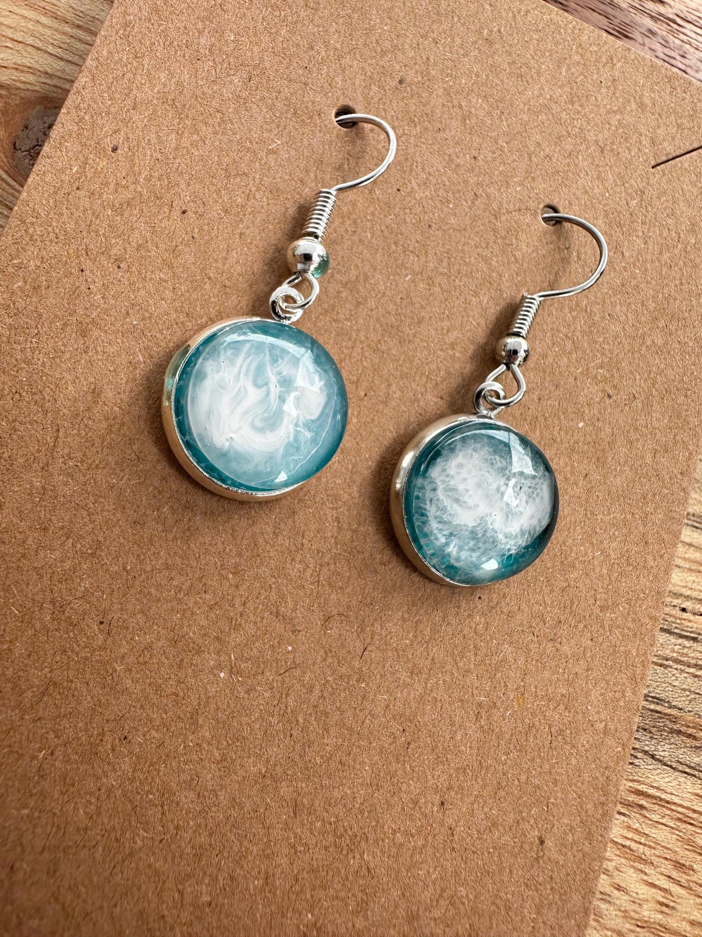 A pair of resin earrings featuring swirling shades of blue and green, resembling a stormy sea. The earrings are glossy, with silver hooks and a unique wave pattern in each piece. Set against a neutral background to highlight the ocean-inspired design