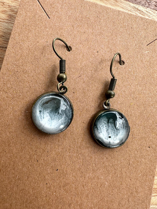 A pair of resin earrings featuring swirling shades of blue and green, resembling a stormy sea. The earrings are glossy, with silver hooks and a unique wave pattern in each piece. Set against a neutral background to highlight the ocean-inspired design
