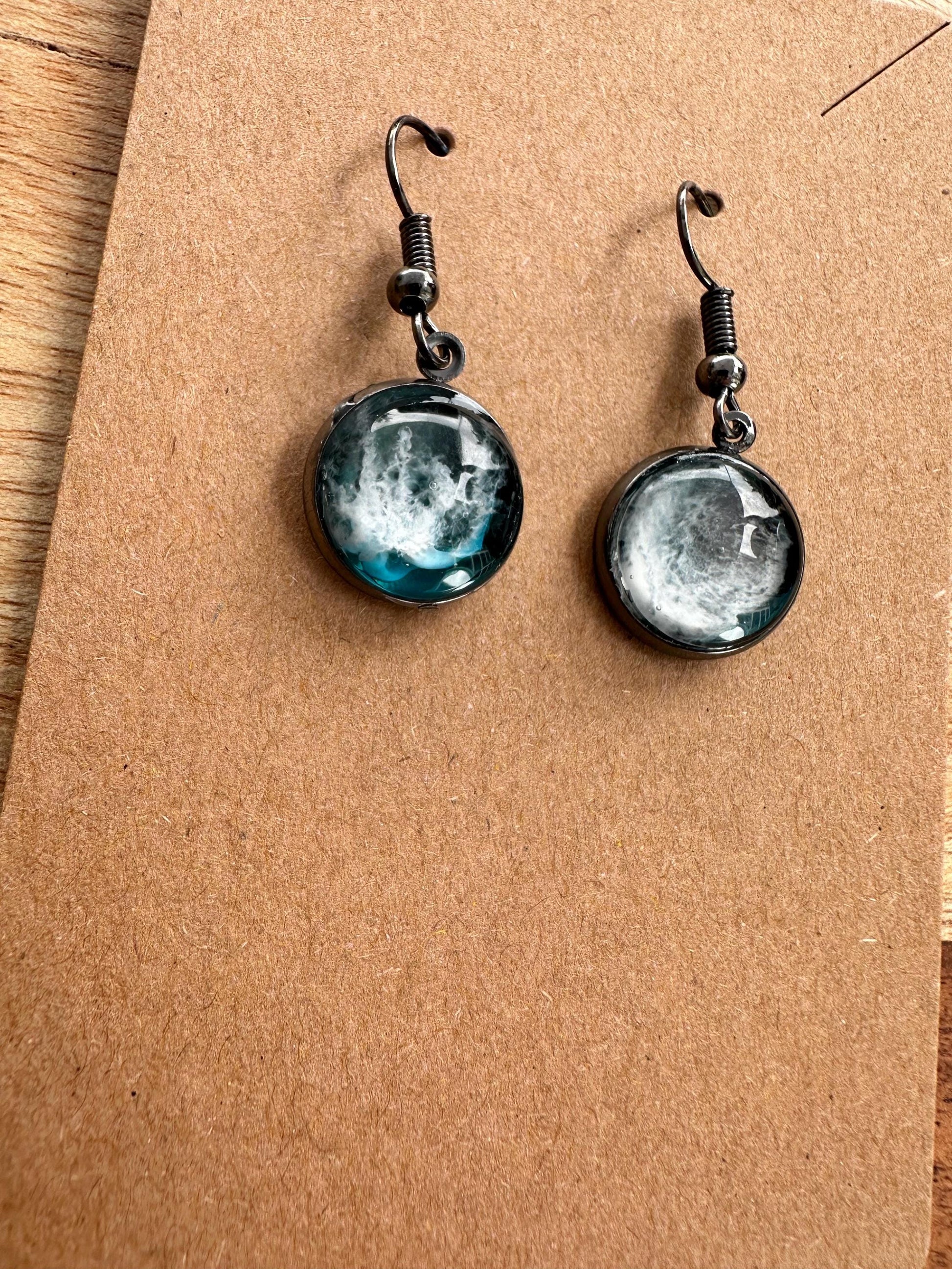 A pair of resin earrings featuring swirling shades of blue and green, resembling a stormy sea. The earrings are glossy, with silver hooks and a unique wave pattern in each piece. Set against a neutral background to highlight the ocean-inspired design