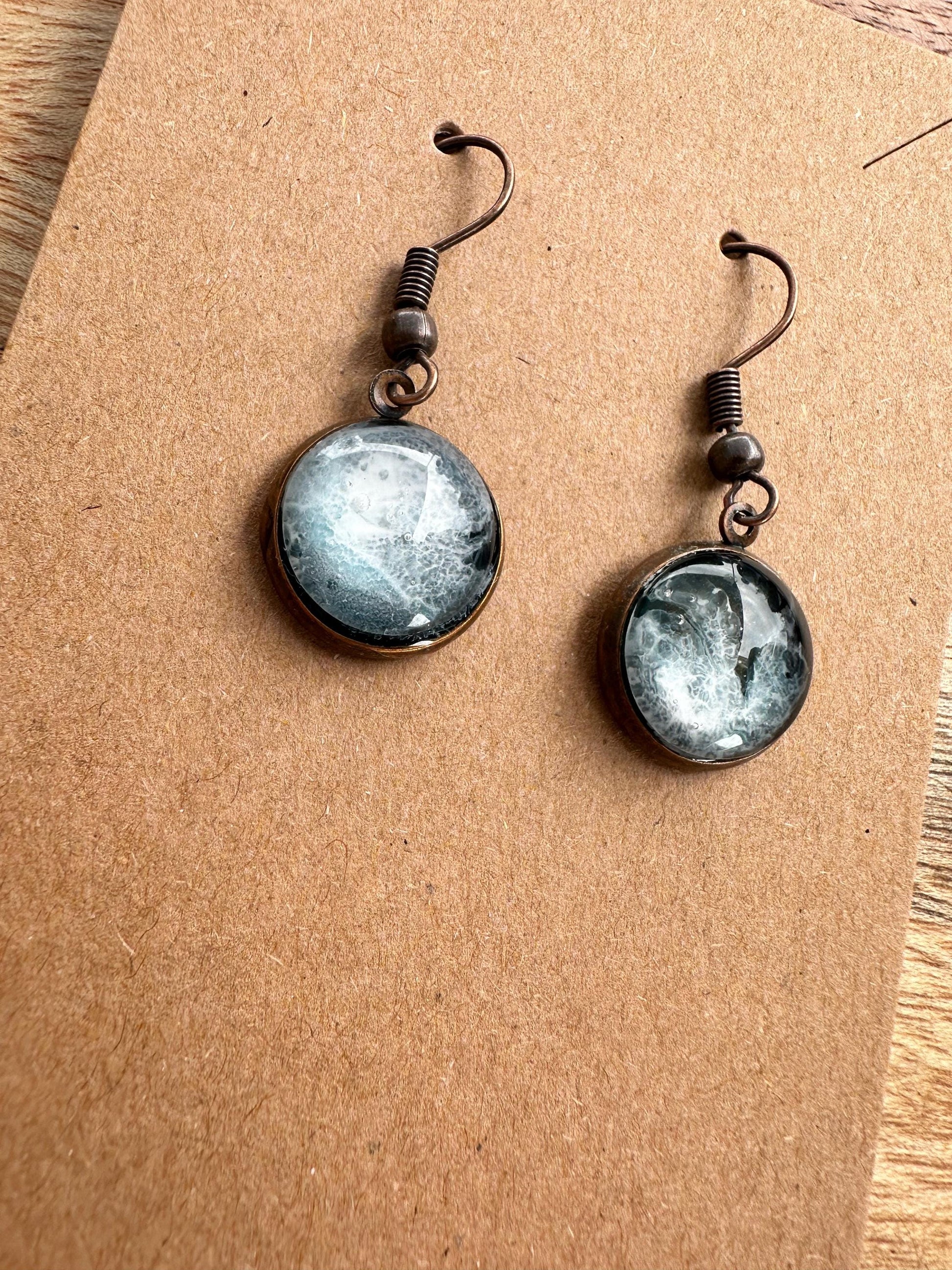 A pair of resin earrings featuring swirling shades of blue and green, resembling a stormy sea. The earrings are glossy, with silver hooks and a unique wave pattern in each piece. Set against a neutral background to highlight the ocean-inspired design