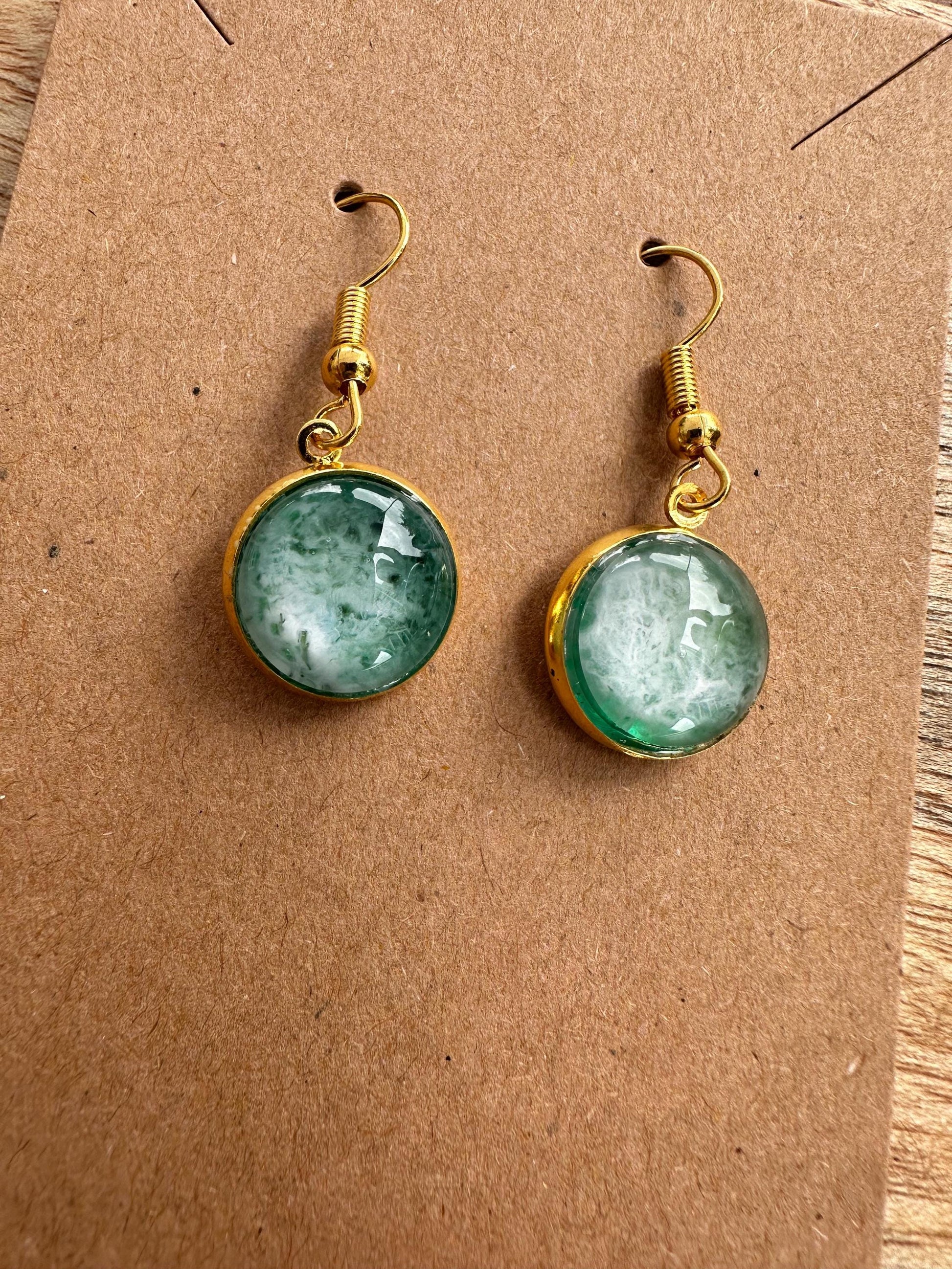 A pair of resin earrings featuring swirling shades of blue and green, resembling a stormy sea. The earrings are glossy, with silver hooks and a unique wave pattern in each piece. Set against a neutral background to highlight the ocean-inspired design
