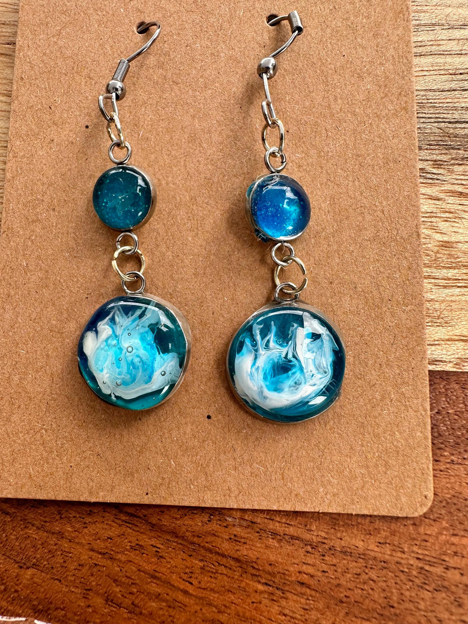 A pair of resin earrings featuring swirling shades of blue and green, resembling a stormy sea. The earrings are glossy, with silver hooks and a unique wave pattern in each piece. Set against a neutral background to highlight the ocean-inspired design